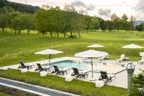 Zona junto a la piscina con sillones de descanso y sombrillas, rodeada de un campo de césped y árboles.