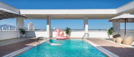 A rooftop pool with a clear view of the city skyline, a pink flamingo float, and a covered seating area with umbrellas.