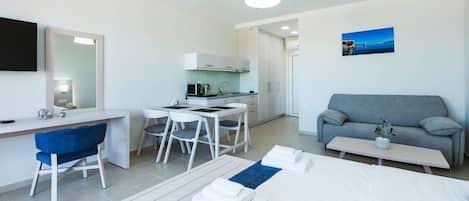 A modern hotel room with a bed, a desk with a chair, a sofa, a small table, and a kitchen area.