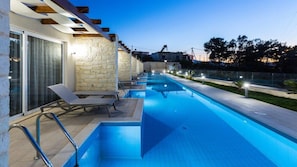 A swimming pool with a stone wall and a glass door, surrounded by lounge chairs and a paved deck.