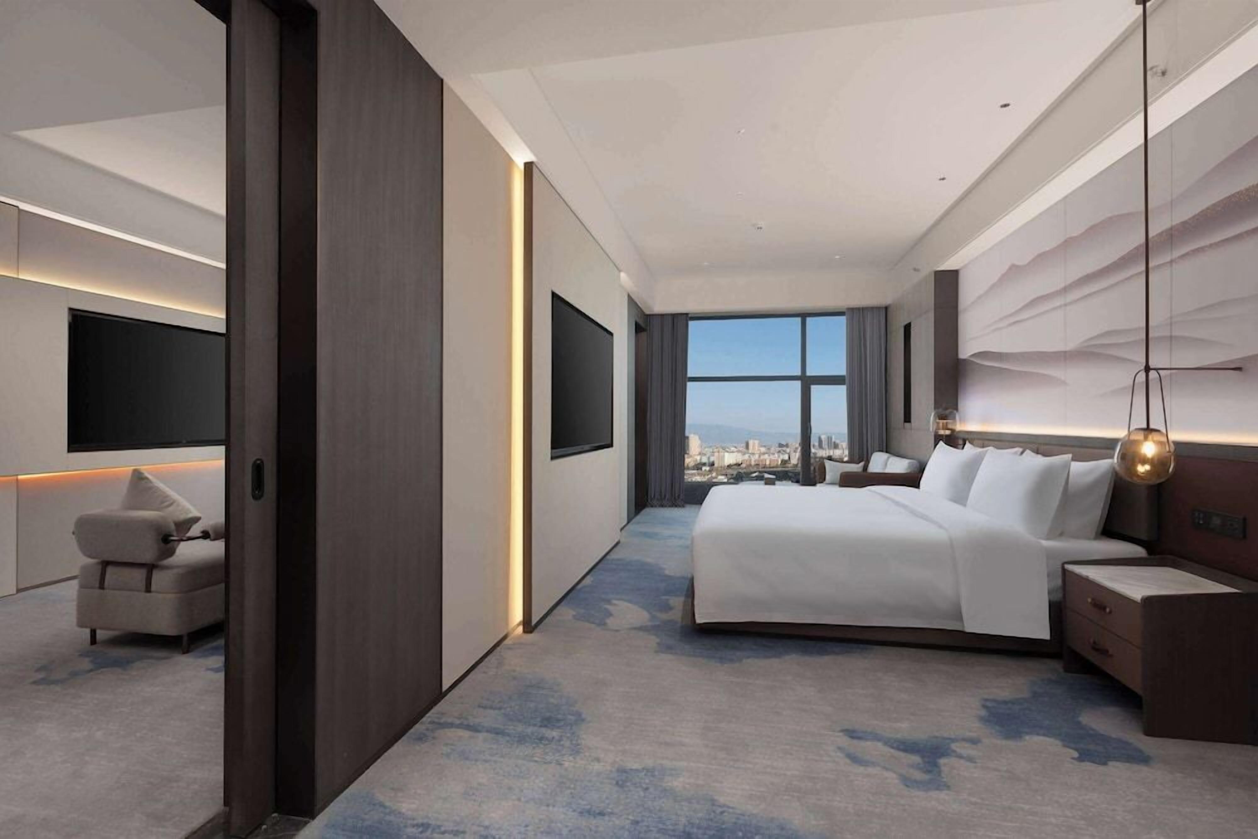 Quarto de hotel moderno com cama grande, sofá, televisão e vista para a cidade.