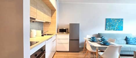 Eine moderne Küche mit weißen Schränken, einem Backofen und einem Kühlschrank. Es gibt eine Essecke mit Tisch und Stühlen, und eine Couch im Hintergrund.