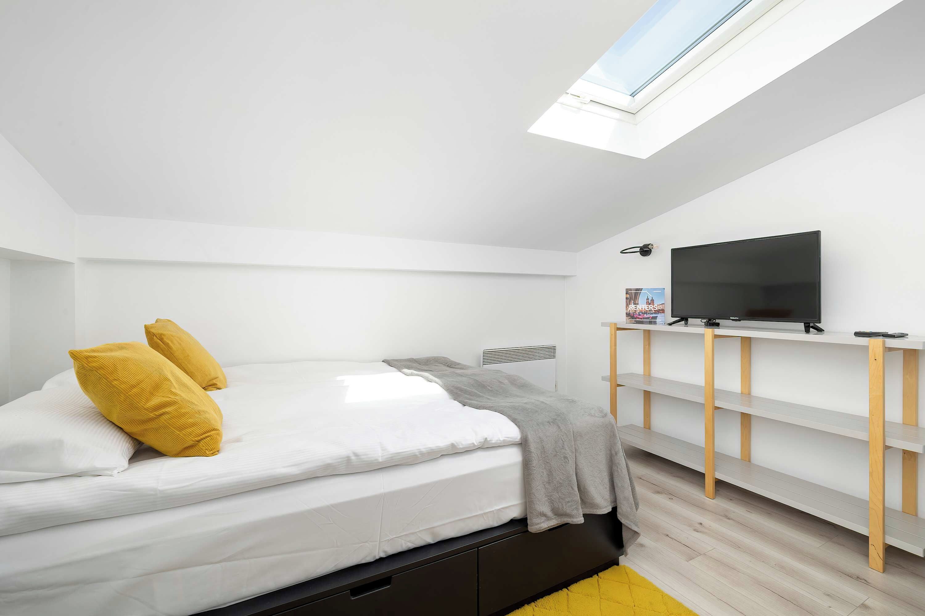 Ein modernes Schlafzimmer mit Oberlicht, einem Bett mit weißer Bettwäsche und zwei gelben Kissen, einer grauen Tagesdecke, einem Fernseher auf einem Holzregal und einem Heizkörper.