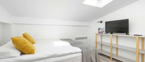 A modern bedroom with a skylight, a bed with white bedding and two yellow pillows, a gray throw blanket, a television on a wooden shelf, and a radiator.
