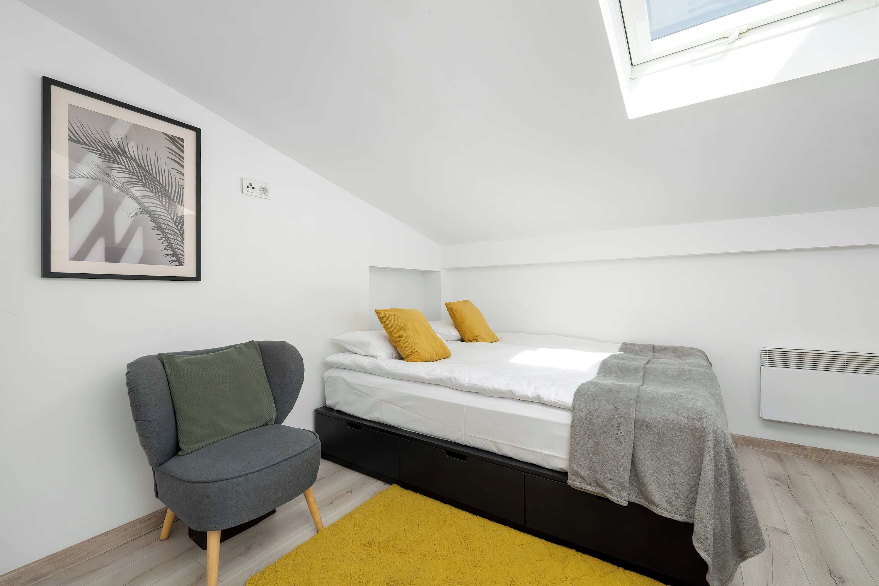 Ein kleines, modernes Schlafzimmer mit einem Bett, einem Sessel, einem Heizkörper und einem gerahmten Bild an der Wand.