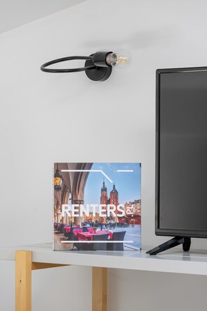 A flat-screen monitor, a wall-mounted lamp, and a book titled 'RENTERS' on a white shelf. - Cracow | 11 Mazowiecka Street | Cosy Studio | Air conditioning (Kraków)
