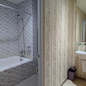Un baño con ducha revestida de azulejos, zona de ducha con cerramiento de vidrio, lavabo con espejo y cubo de basura.