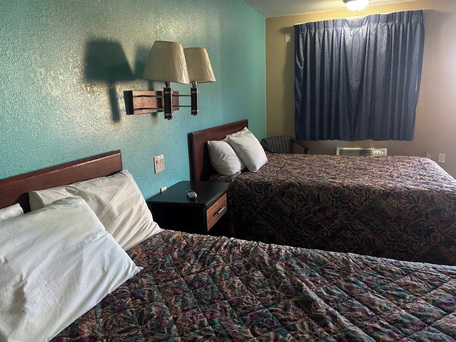 Une chambre d’hôtel avec deux lits, une table de chevet, une lampe et une fenêtre avec des rideaux.