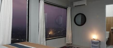 A hotel room with a bed, a nightstand, a mirror, and a view of the city at night.