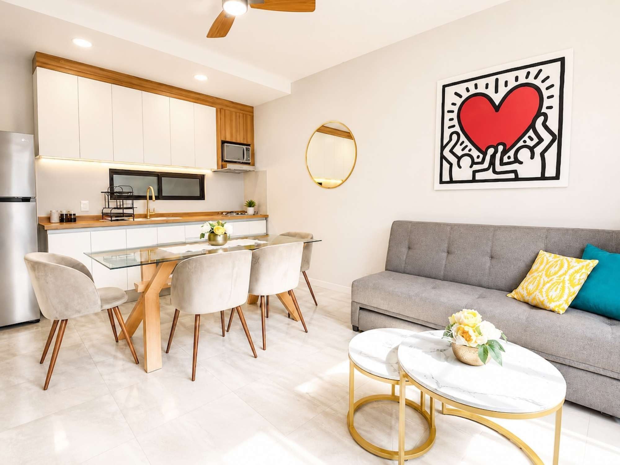 A modern kitchen with white cabinets, a dining area with a glass table, and a grey sofa with yellow pillows.