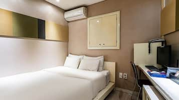 A hotel room with a bed, a desk with a computer, an air conditioning unit, and a wall-mounted TV.
