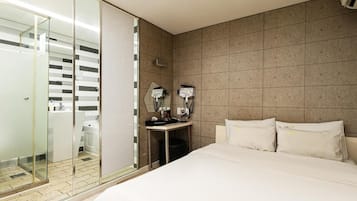 A hotel room with a large bed, a glass-enclosed shower area, a small bedside table, and a wall-mounted light fixture.