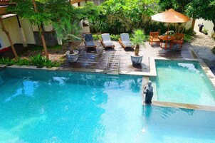 Una piscina con sillones de descanso y una pequeña mesa, rodeada de plantas en macetas y una terraza de madera.