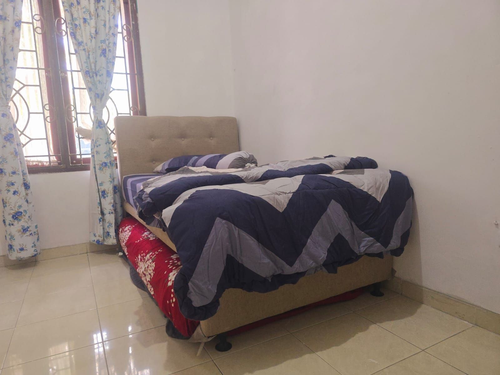 Uma cama de solteiro com cabeceira, um travesseiro vermelho e um cobre-leito estampado em um quarto com piso de azulejos e janela com cortinas.