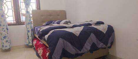 Uma cama de solteiro com cabeceira, um travesseiro vermelho e um cobre-leito estampado em um quarto com piso de azulejos e janela com cortinas.
