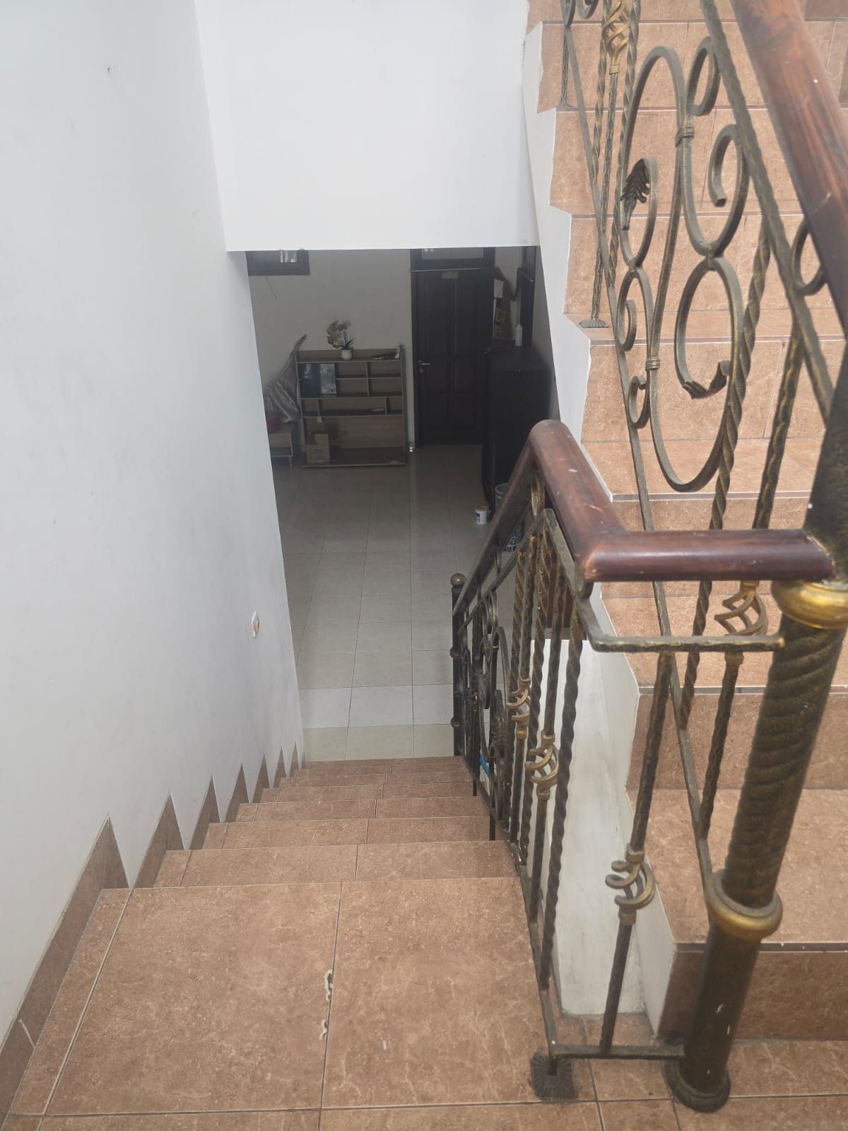 Una escalera con barandilla de metal y pasamanos de madera, que conduce a un espacio interior con piso de baldosas y una puerta.