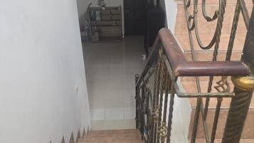 Una escalera con barandilla de metal y pasamanos de madera, que conduce a un espacio interior con piso de baldosas y una puerta.