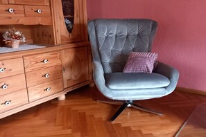 A room with a tufted grey armchair with a single cushion, a wooden cabinet with drawers, and a herringbone-patterned wooden floor. - Holiday apartment with 2 bedrooms (Geiselwind)