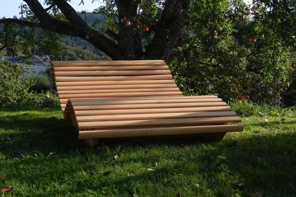 A wooden bench with horizontal slats, situated on a grassy area with trees and a hill in the background.