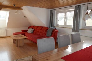 A modern living room with a red sofa, grey chairs, a wooden dining table, and a window with curtains.