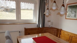 Una habitación con una mesa de madera, un camino de mesa rojo, una ventana con cortinas, un radiador y una lámpara.