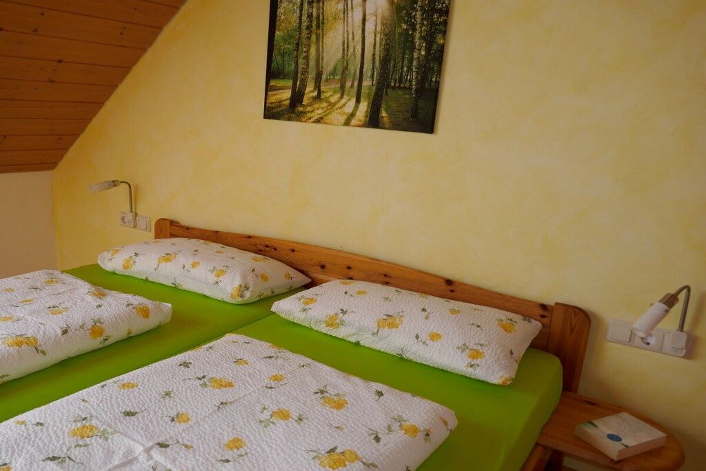 A room with a wooden sloped ceiling, a bed with green bedding and floral-patterned pillows, a wall-mounted lamp, and a painting of a forest.