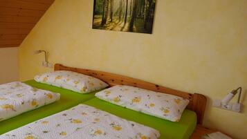 A room with a wooden sloped ceiling, a bed with green bedding and floral-patterned pillows, a wall-mounted lamp, and a painting of a forest.