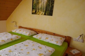 A room with a wooden sloped ceiling, a bed with green bedding and floral-patterned pillows, a wall-mounted lamp, and a painting of a forest.