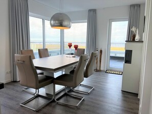 A modern dining area with a rectangular table, six chairs, a pendant light, and a view of the sea through large windows. - Holiday Apartment With 1 Bedroom (Wangerland)