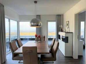 A modern dining area with a large table and chairs, a pendant light, and a view of the beach through large windows. - Holiday Apartment With 1 Bedroom (Wangerland)