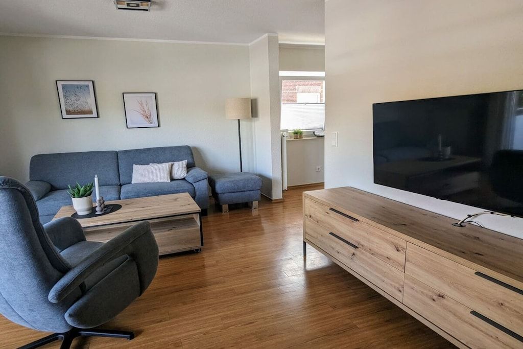 Un salon moderne avec un canapé gris, une table basse en bois, un fauteuil gris et un téléviseur posé sur un meuble en bois.