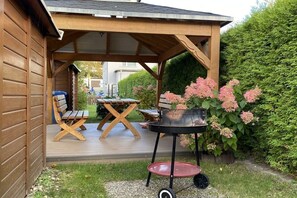 Un patio in legno con zona salotto coperta, un barbecue e un vaso di fiori.