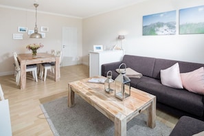 A living room with a wooden coffee table, a grey sofa, and a dining area with a wooden table and chairs. - Kaiserhof 002 Reinke am Strand (Norderney)