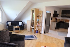 A modern living room with a dark sofa, a small table, and a bookshelf. A kitchen with a refrigerator and a TV is visible in the background. - Holiday apartment in Süsel with swimming pool (Süsel)