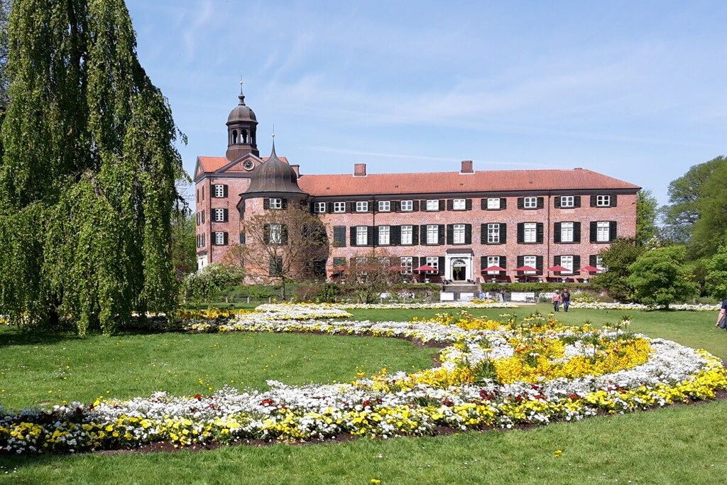 A large building with a prominent tower and a symmetrical facade, surrounded by a well-maintained garden with flower beds.