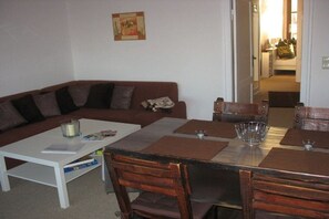 Un espace de vie comprenant un canapé, une table à manger et des chaises. Un tableau est accroché au mur, et une porte donne accès à une autre pièce.