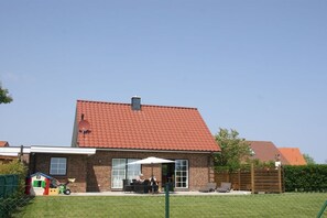 Ein zweistöckiges Backsteinhäuschen mit rotem Ziegeldach, einer Terrasse mit Gartenmöbeln und einem umzäunten Hof.