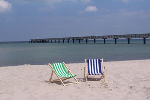 Zwei Strandstühle an einem sandigen Strand, im Hintergrund ein Steg.