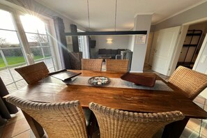 A dining area with a wooden table and wicker chairs, a view of the outdoors through a glass door, and a living area in the background. - Jona with sauna/fireplace (Schönberg)