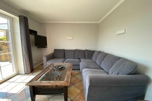 Ein Wohnzimmer mit grauem Ecksofa, einem Glastisch, einem am Wand montierten Flachbildfernseher und einer Glastür, die zu einem Außenbereich führt.