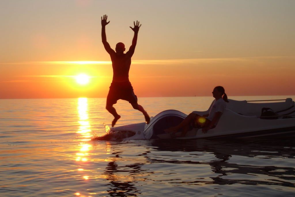 A person standing on a boat with arms raised, another person sitting on the boat, and the sun setting in the background.