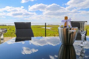 Ein Tisch mit einer Champagnerflasche und einem Glas, vor einer Aussicht auf ein grünes Feld und einen klaren Himmel.
