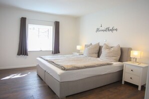 Une chambre à coucher avec un lit, deux tables de chevet, une fenêtre avec des rideaux et un mur orné d’une affiche.