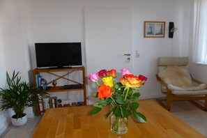 Una sala de estar con un televisor sobre un mueble bajo, una mesa de madera con un florero, una silla y una estantería.