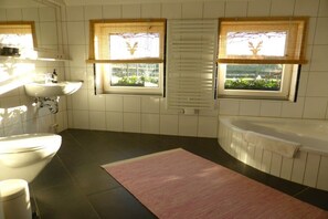 Un baño con lavabo, inodoro y bañera. Hay ventanas con persianas y vistas al exterior con vegetación.