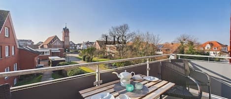 Un balcon avec une table dressée pour le thé, donnant sur une ville dont on aperçoit le clocher d’une église.