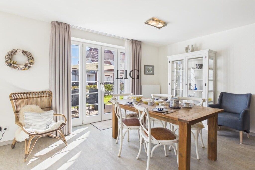 Un coin repas avec une table en bois, des chaises blanches, un fauteuil en osier, un buffet et une vue à travers une porte vitrée.
