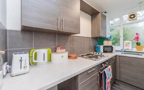 Uma cozinha moderna com armários cinza, bancada branca, torradeira, chaleira, micro-ondas e janela com vista para o verde.