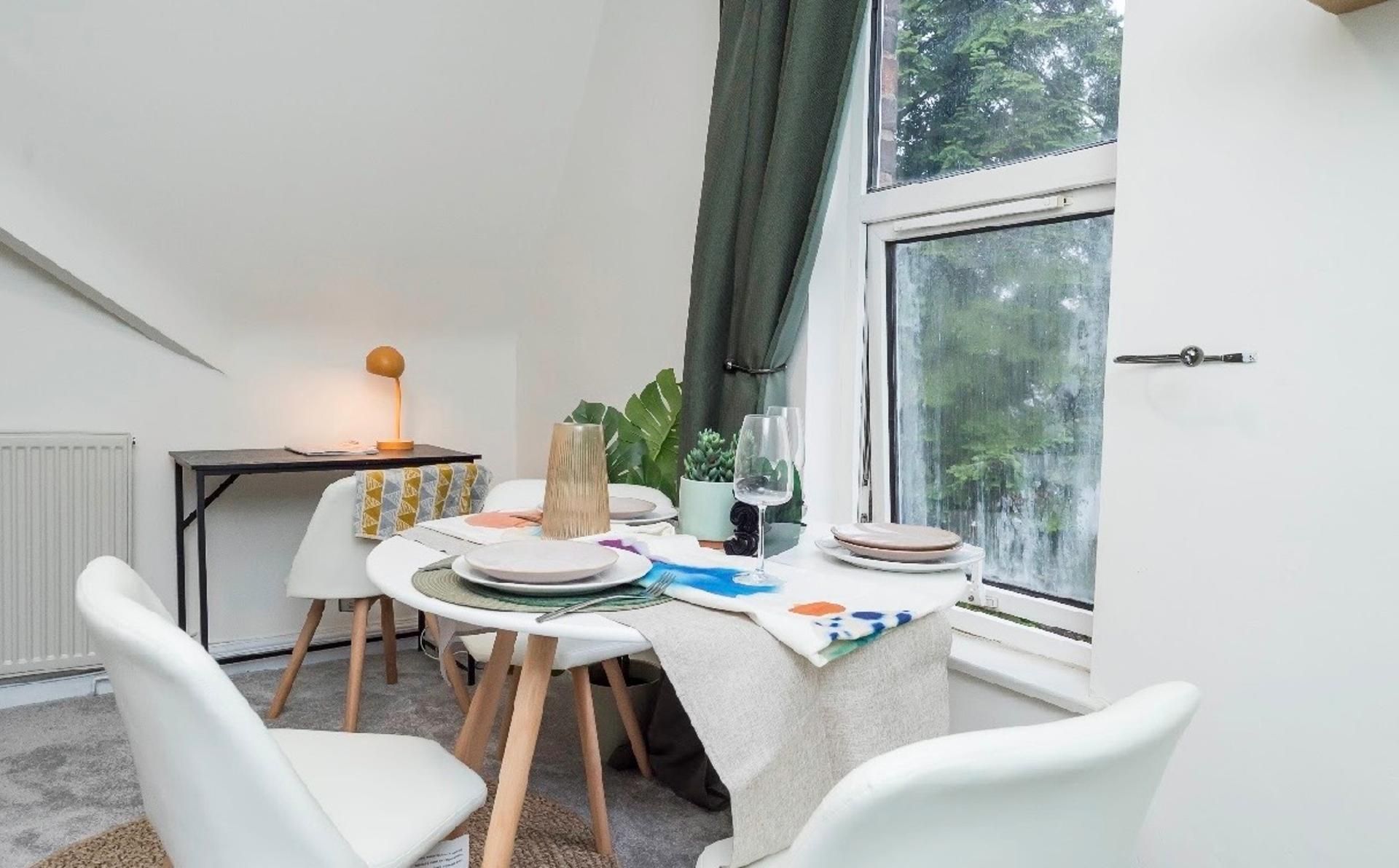 Uma área de refeições com mesa posta para dois, uma luminária e vista para o exterior pela janela.