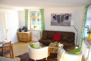A living room with a brown sofa, two white armchairs, a coffee table, a side table with speakers, a window with green curtains, and a painting on the wall. - Holiday apartment with 2 bedrooms (Wahlstorf-Wielen)
